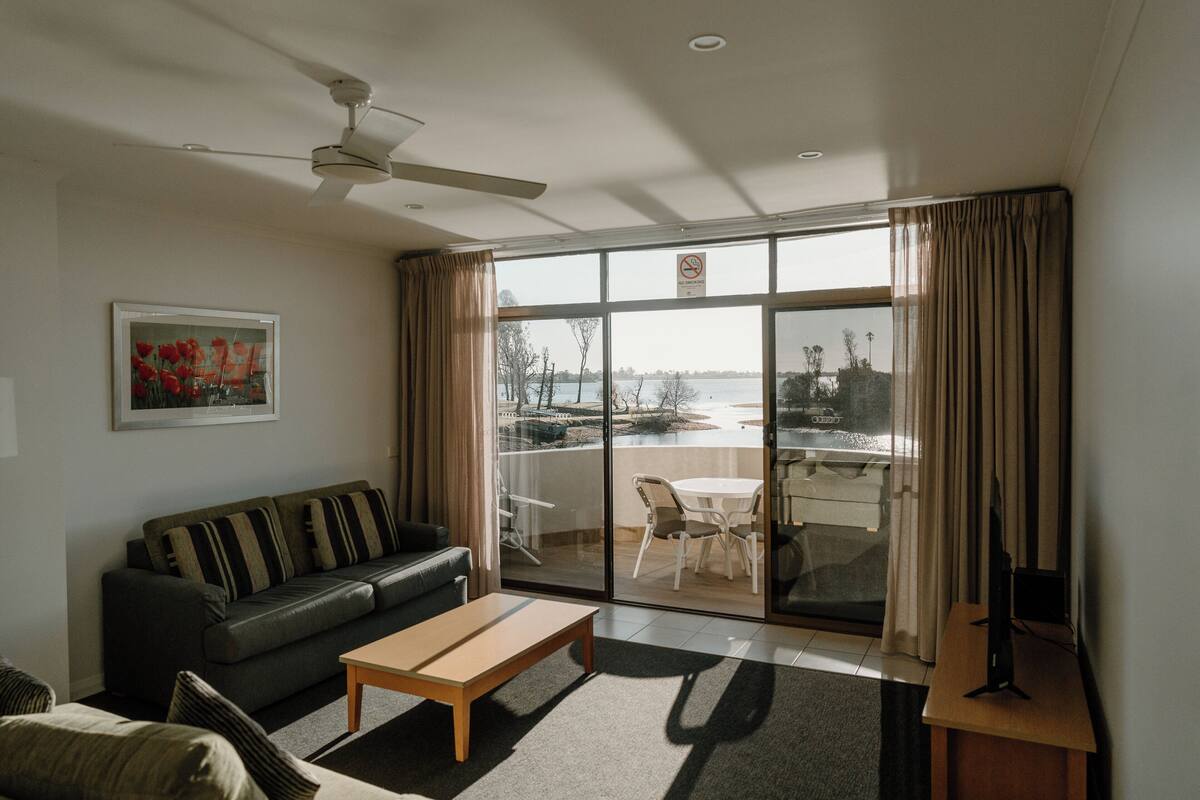 A living room with a sofa, a coffee table, and a TV stand. There is a large window with a view of a body of water and a boat.
