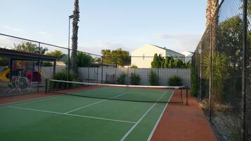 An outdoor tennis court with a net, surrounded by a chain-link fence, and adjacent to a children's play area.