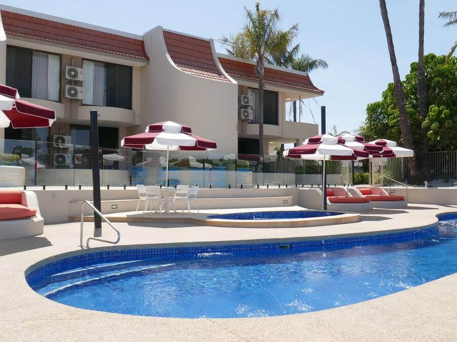 A swimming pool with red and white sun loungers and umbrellas, surrounded by a building with a tiled roof and air conditioning units.