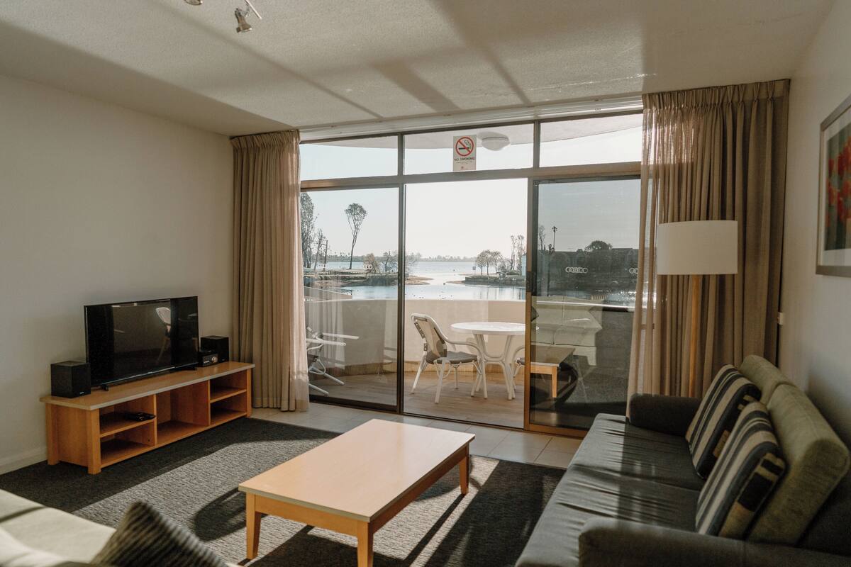 A hotel room with a television on a wooden stand, a sofa, a table, and a balcony with a view of the sea.
