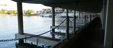 A covered dock with a chain barrier, calm water, and a row of buildings in the background.