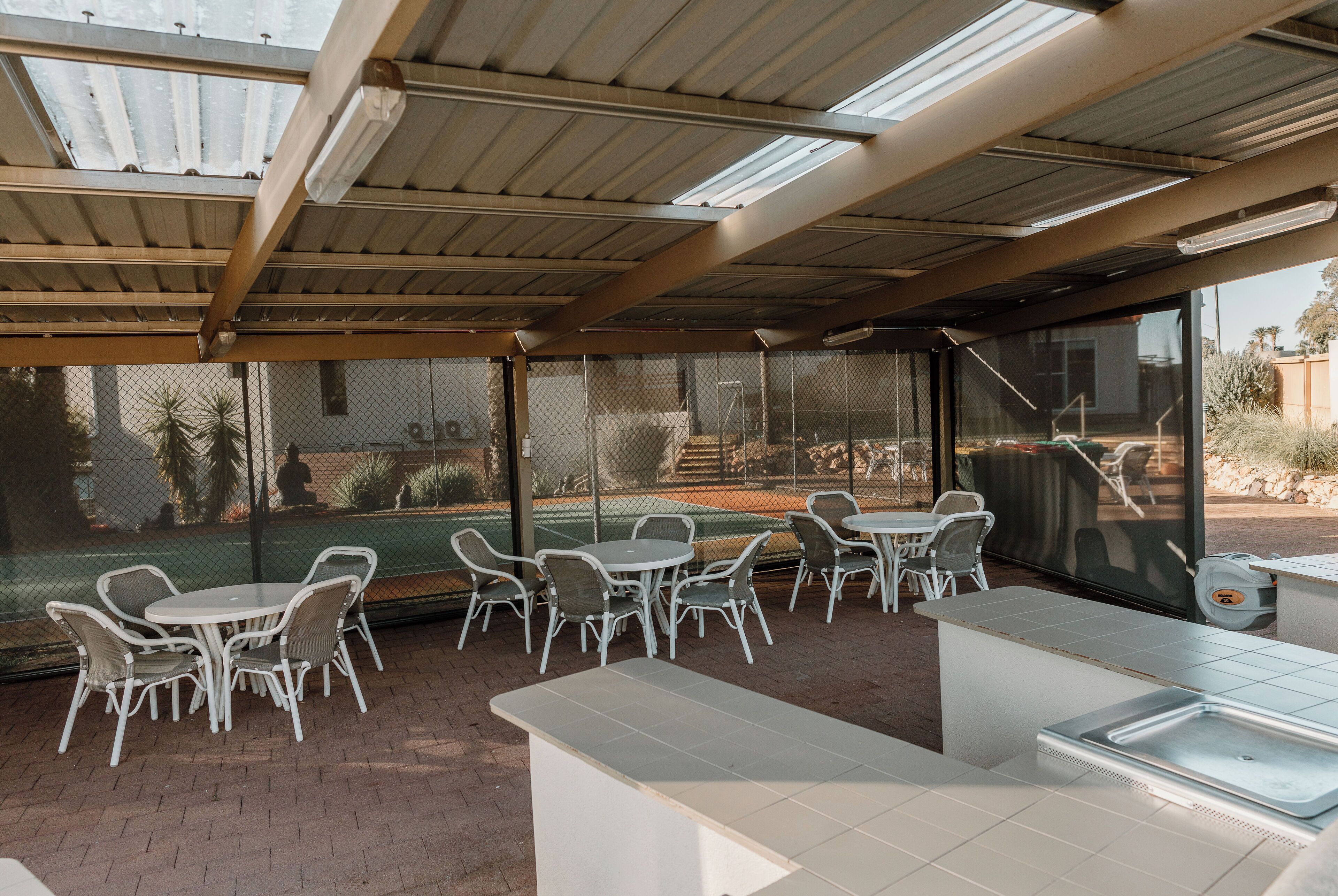 An outdoor patio area with tables and chairs, a covered section with a roof, and a view of a tennis court through a fence.