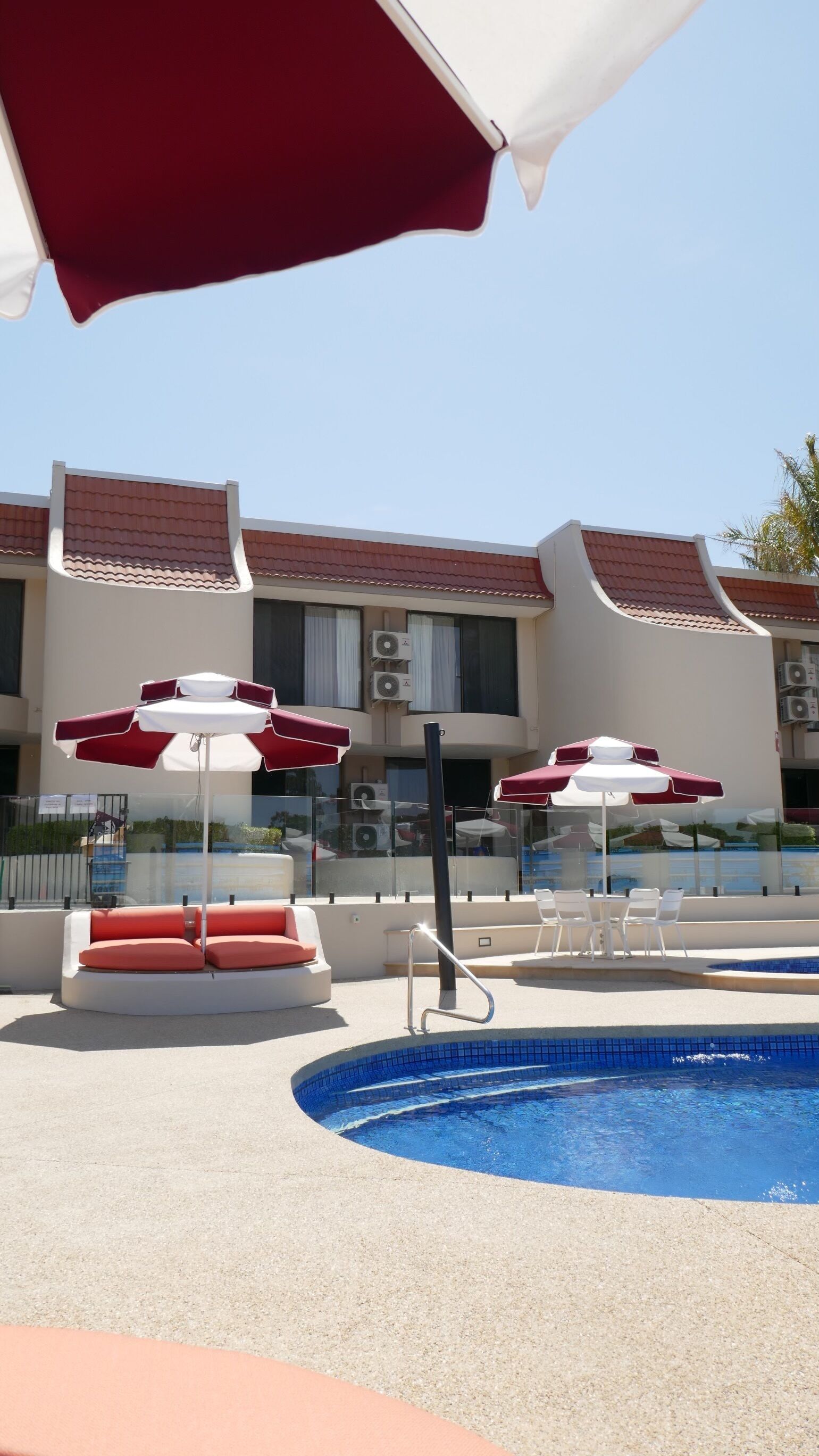 A swimming pool area with red and white umbrellas, lounge chairs, and a building with a tiled roof in the background.