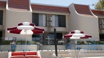 A swimming pool area with red and white umbrellas, lounge chairs, and a building with a tiled roof in the background.