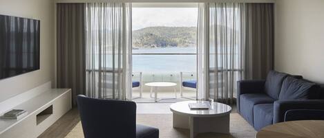 Un moderno salón con vistas a un lago y colinas, que incluye un sofá, una mesa de centro y un televisor sobre un mueble.