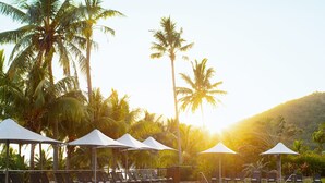 A pool area with lounge chairs and umbrellas, surrounded by palm trees and a mountain in the background.