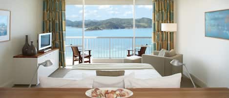 Ein Hotelzimmer mit einem großen Bett, einem Schreibtisch mit Obstschale, einem Fernseher und Blick auf das Meer.