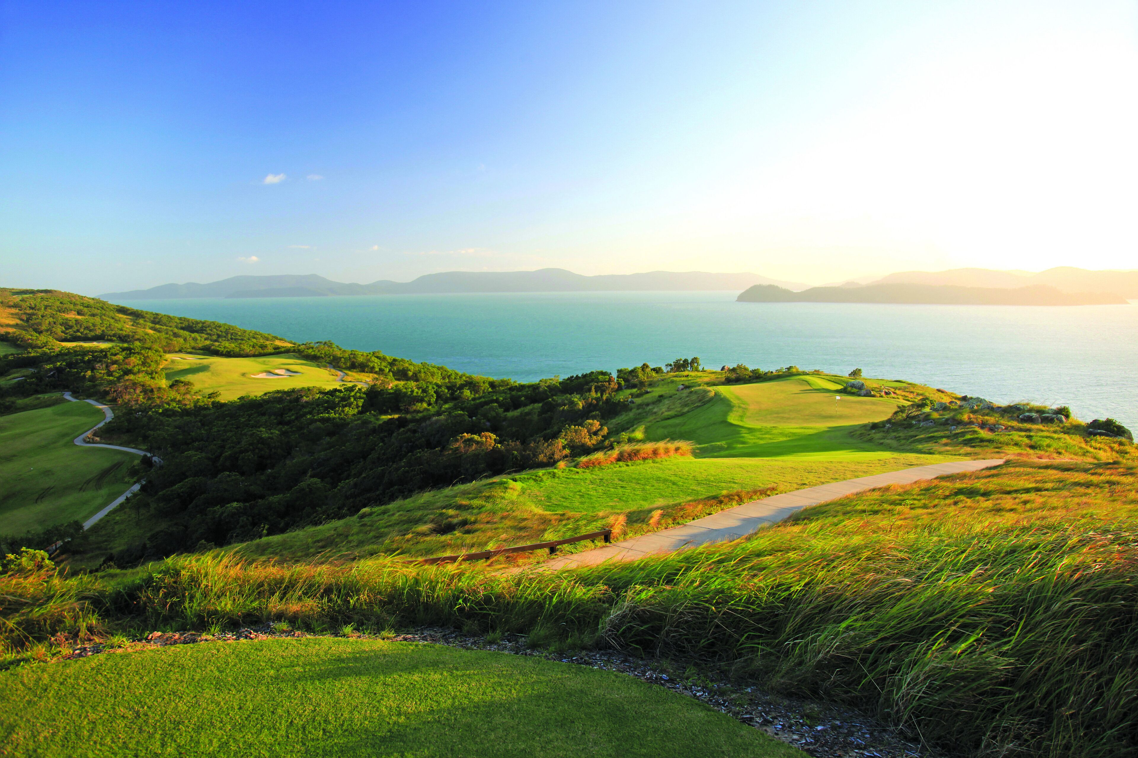 A golf course with a path leading to the water, surrounded by green hills and a clear sky.
