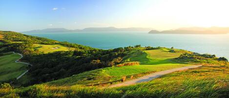 A golf course with a path leading to the water, surrounded by green hills and a clear sky.