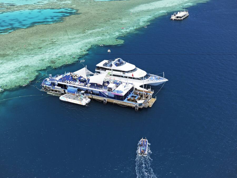 An aerial view of a floating structure with multiple decks and a large open deck area, surrounded by clear blue waters and a coral reef.