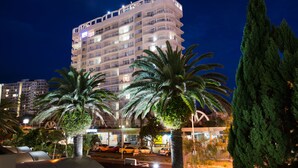 Un hotel a più piani con balconi, palme e un'area di parcheggio.