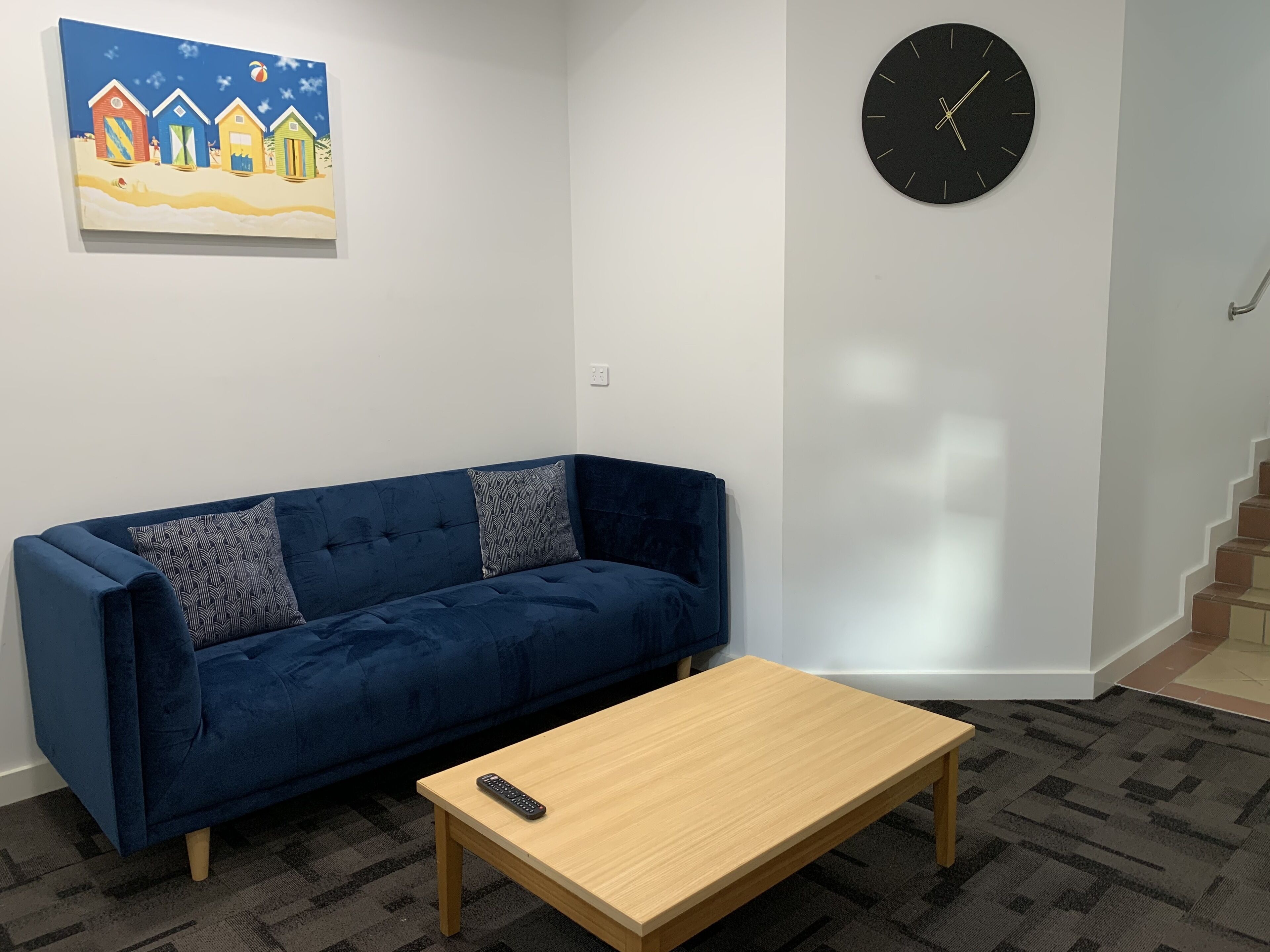 A living room with a blue sofa, a wooden coffee table, a black clock, and a painting of beach huts.