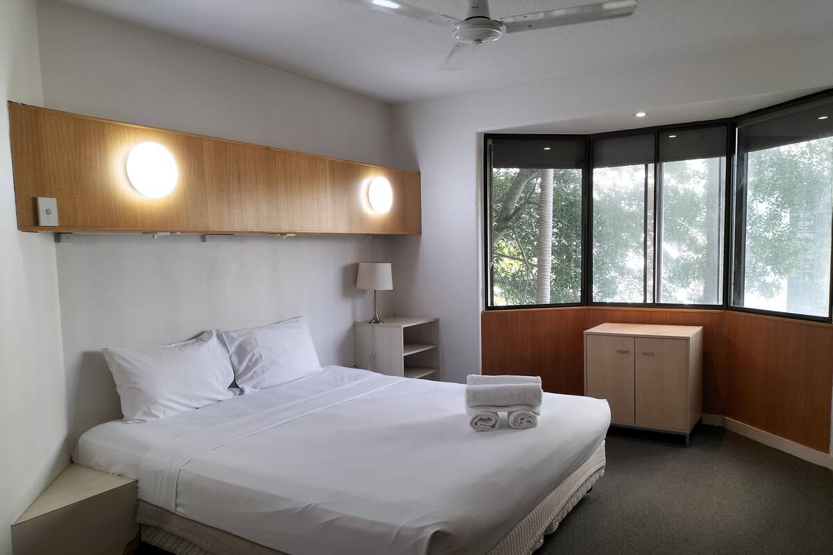 Habitación de hotel con cama, mesita de noche, lámpara y ventana con vistas a árboles.
