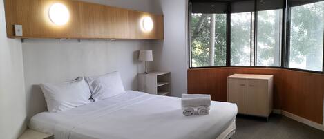 A hotel room with a bed, bedside table, lamp, and a window with a view of trees.