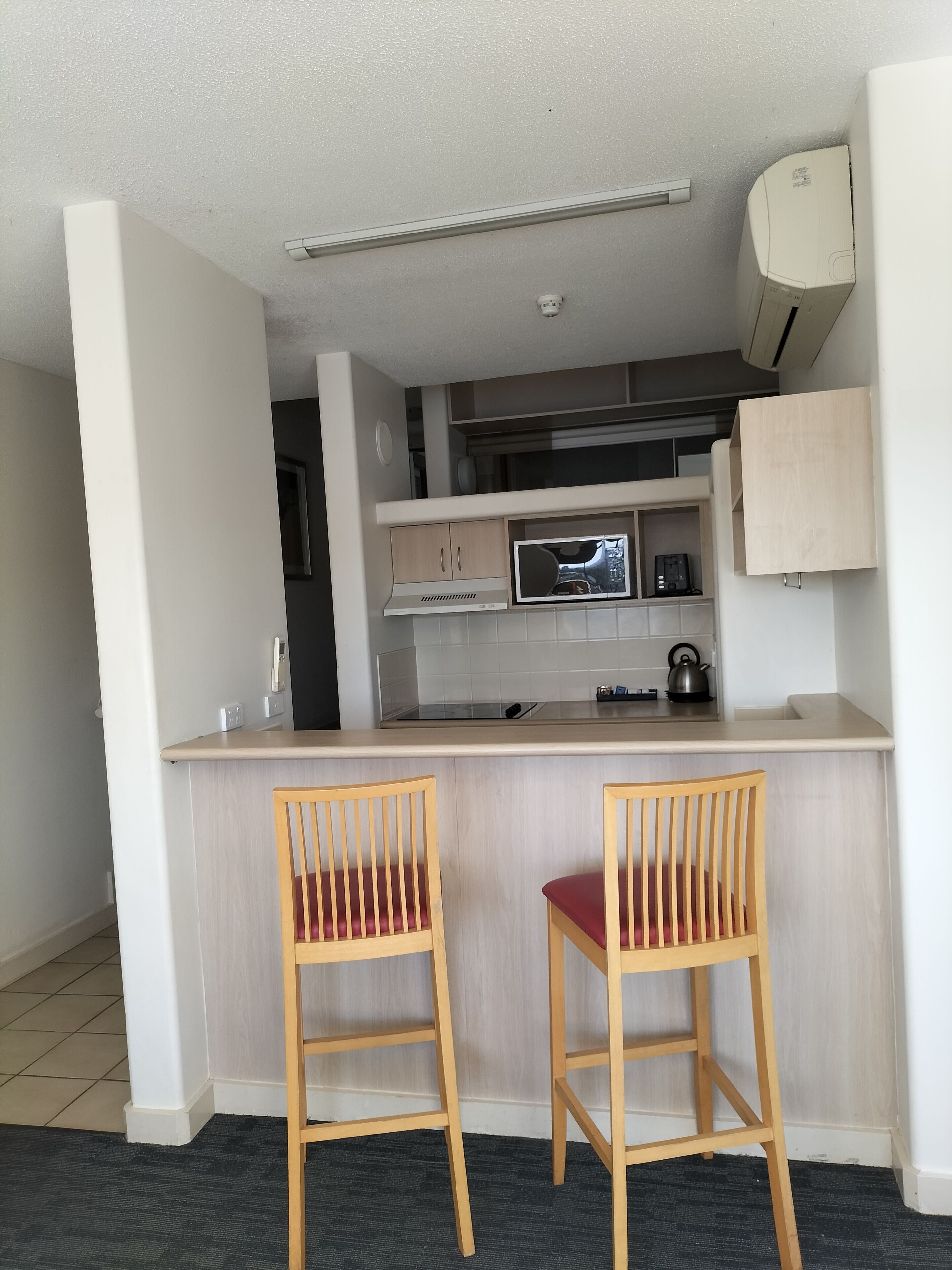 A compact kitchen with a microwave, stove, and cabinets. There are two wooden chairs with red cushions.
