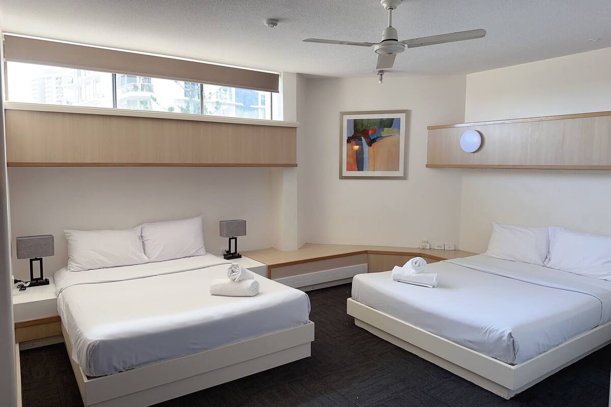 Habitación de hotel con dos camas, ventilador de techo, ventana con persianas, un cuadro y una mesita de noche con lámpara.