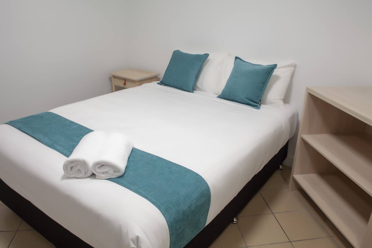 Una cama bien hecha con ropa de cama blanca y turquesa, un par de toallas enrolladas y una mesita de noche pequeña.