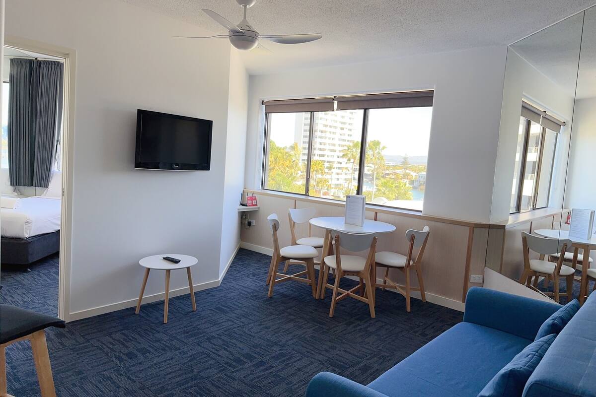 Un cuarto de hotel con una cama, televisión, escritorio con sillas, sofá azul y vistas a edificios y palmeras a través de la ventana.