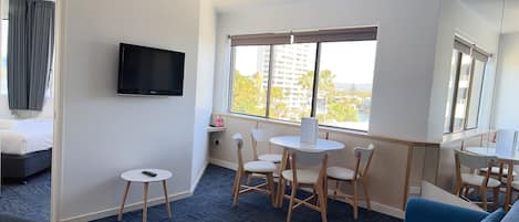 A hotel room with a bed, a TV, a desk with chairs, a blue sofa, and a view of buildings and palm trees through the window.