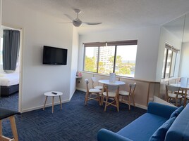 Un cuarto de hotel con una cama, televisión, escritorio con sillas, sofá azul y vistas a edificios y palmeras a través de la ventana.