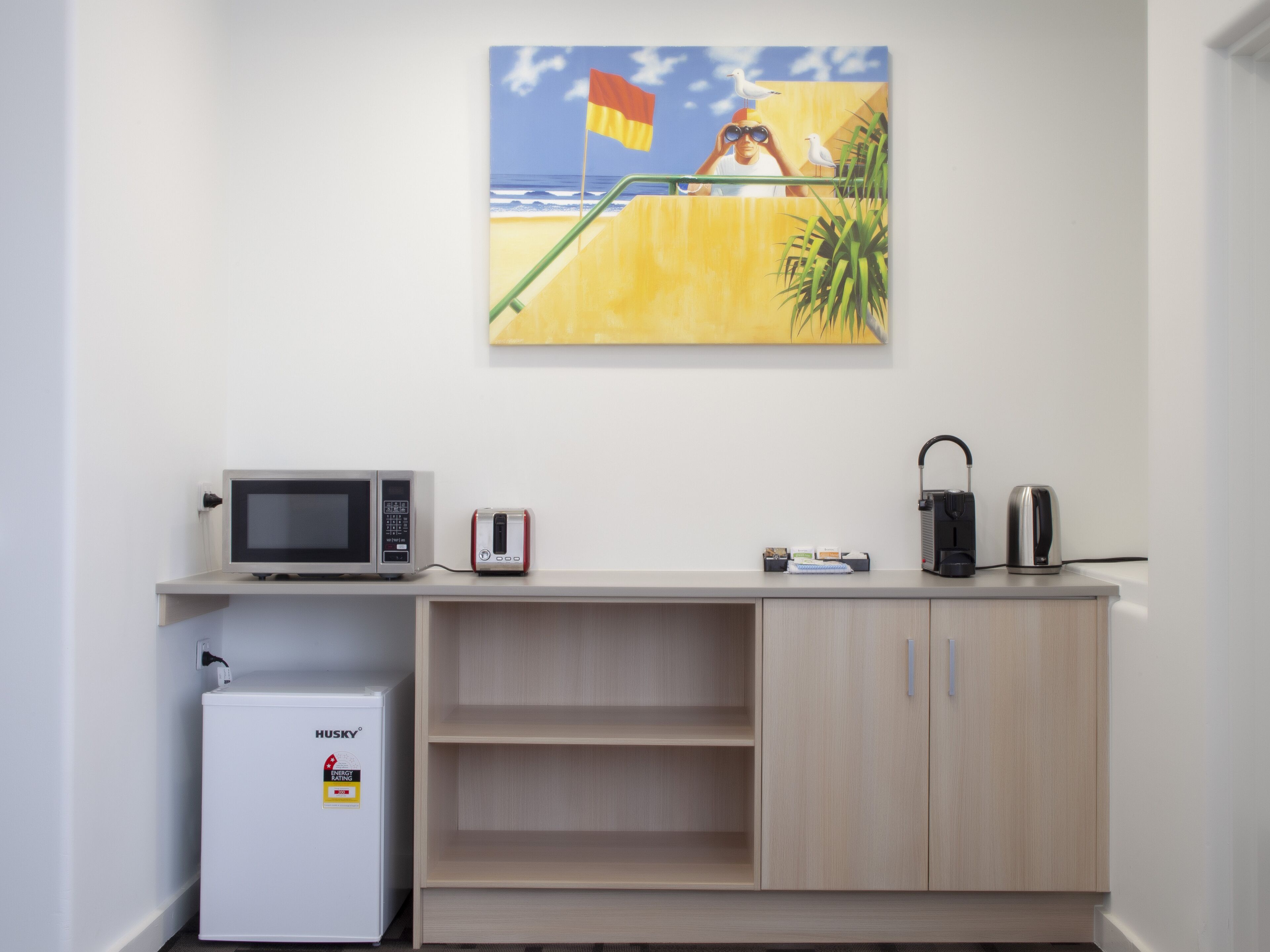 Een kitchenette met een magnetron, broodrooster en koffiezetapparaat op een aanrecht, een kleine koelkast en een houten kast met open planken.