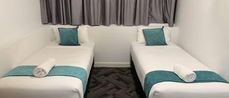 Two single beds with white linens and teal accents, grey curtains, and a patterned carpet.