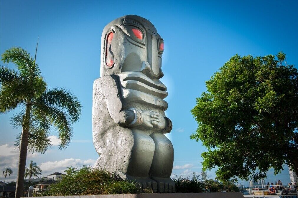 A large stone statue of a figure with a wide mouth and red eyes, standing in an outdoor setting with palm trees and a clear sky.