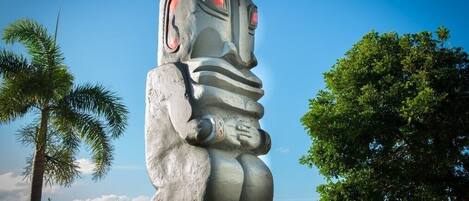 A large stone statue of a figure with a wide mouth and red eyes, standing in an outdoor setting with palm trees and a clear sky.