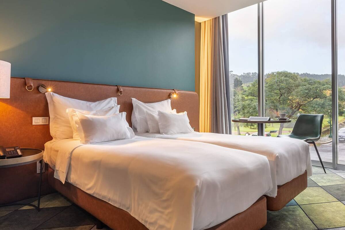 Habitación de hotel con una cama grande, una mesita de noche con lámpara, una pequeña mesa con taza y platillo, y vistas al exterior a través de un ventanal amplio.
