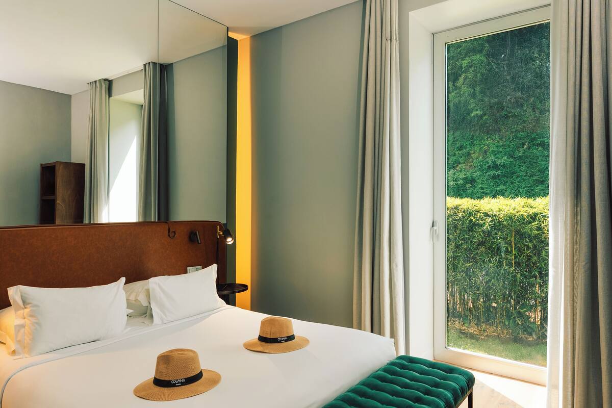 Habitación de hotel con una cama grande, dos sombreros sobre las almohadas, un banco verde, cabecera de madera y vista a la vegetación por la ventana.