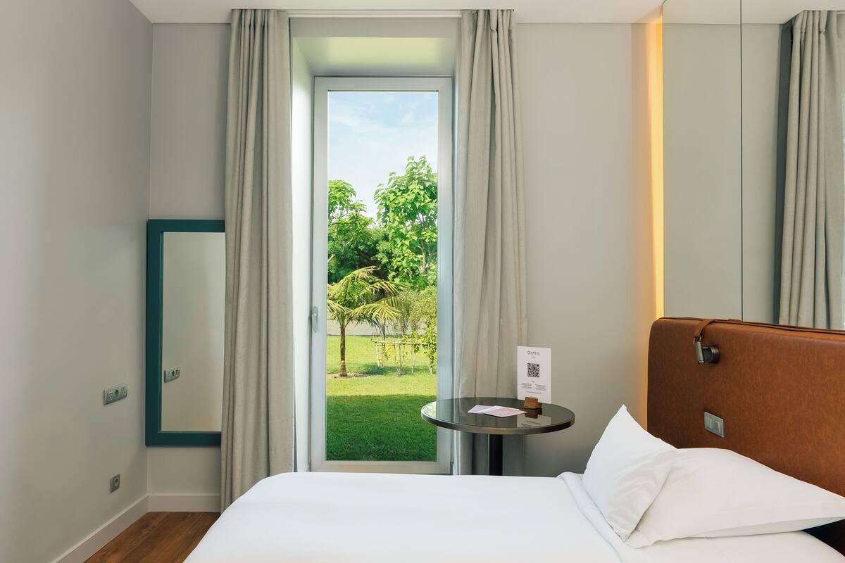 Habitación de hotel con cama, una mesita con un cartel, un espejo y vistas a un jardín a través de una ventana.