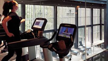 Ein Fitnessstudio mit Laufbändern, auf denen Videos laufen, einer trainierenden Person und Blick ins Freie durch große Fenster.