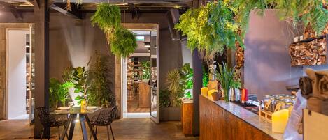 Un interior moderno con un mostrador de recepción de madera, plantas colgantes y un comedor con sillas y una mesa.