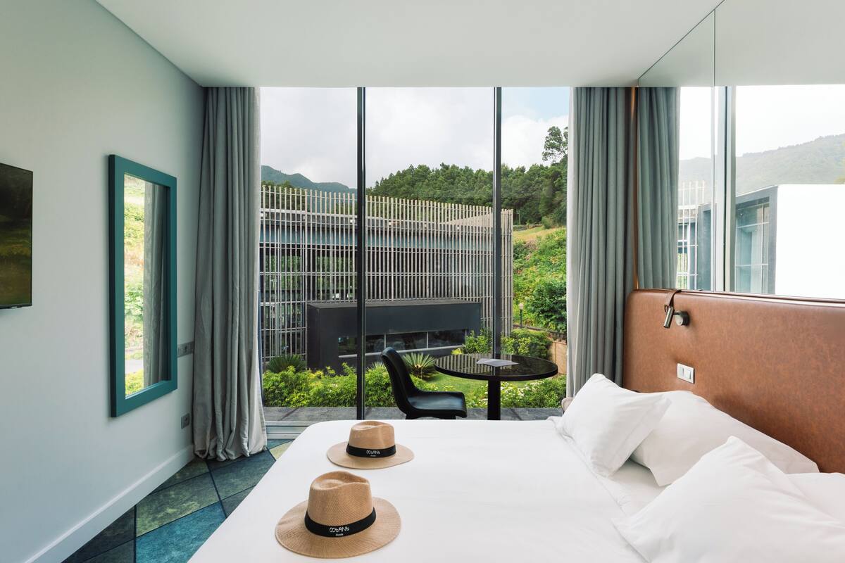 Habitación de hotel moderna con una cama grande, una mesa de comedor y vistas a un paisaje montañoso a través de amplios ventanales.