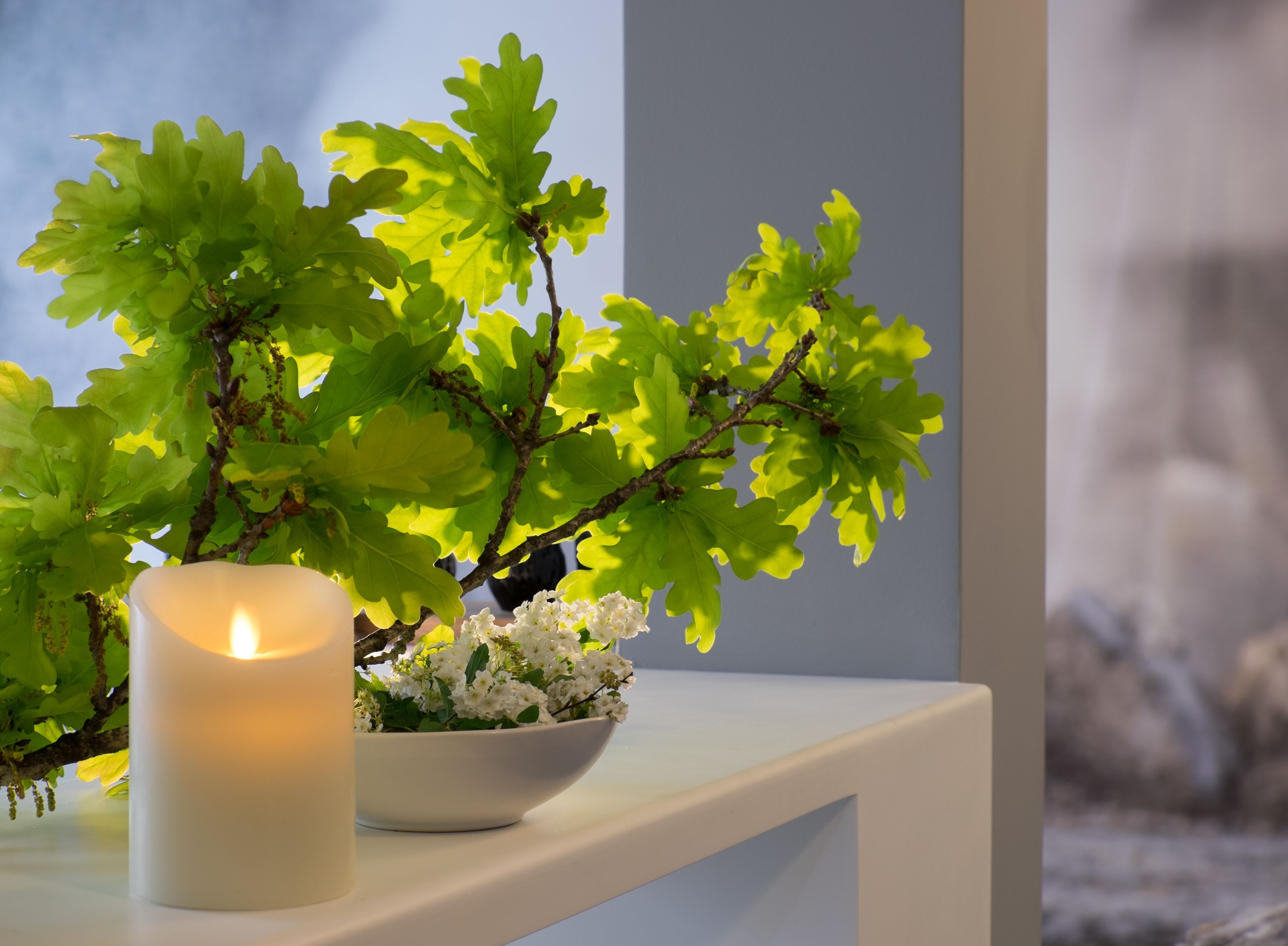 A white candle on a shelf next to a white bowl with white flowers and green leaves.