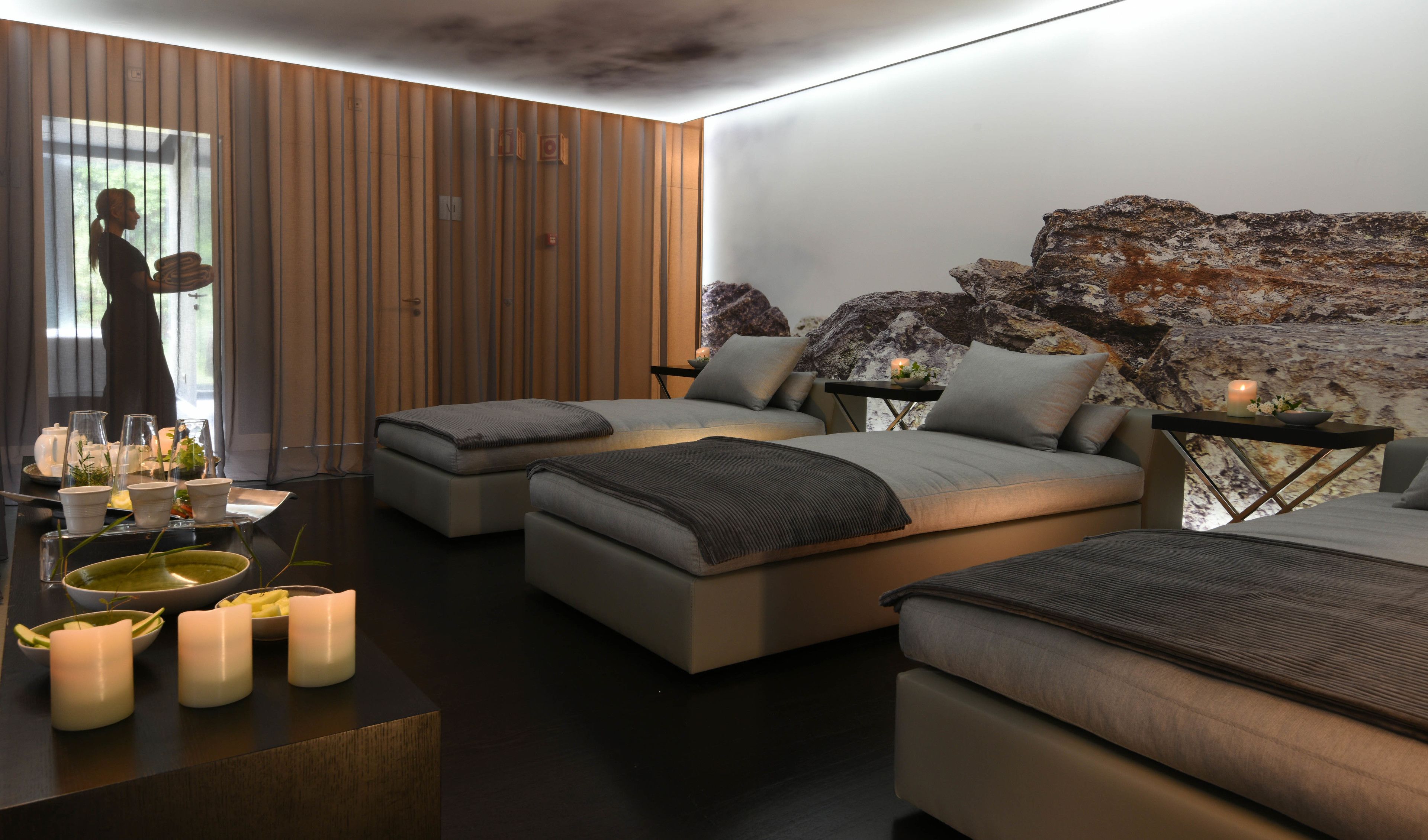 A modern bedroom with a large bed, two smaller beds, a wooden slat wall, a rock wall, and a view of a person outside.