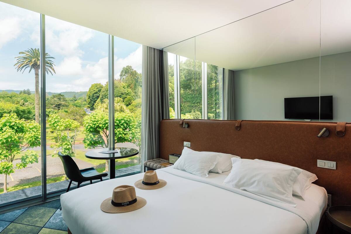 Habitación de hotel con una cama grande, dos sombreros sobre la cama, un televisor en la pared y vistas a zonas verdes al exterior.