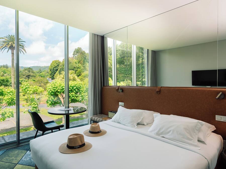 A hotel room with a large bed, two hats on the bed, a TV on the wall, and a view of greenery outside.