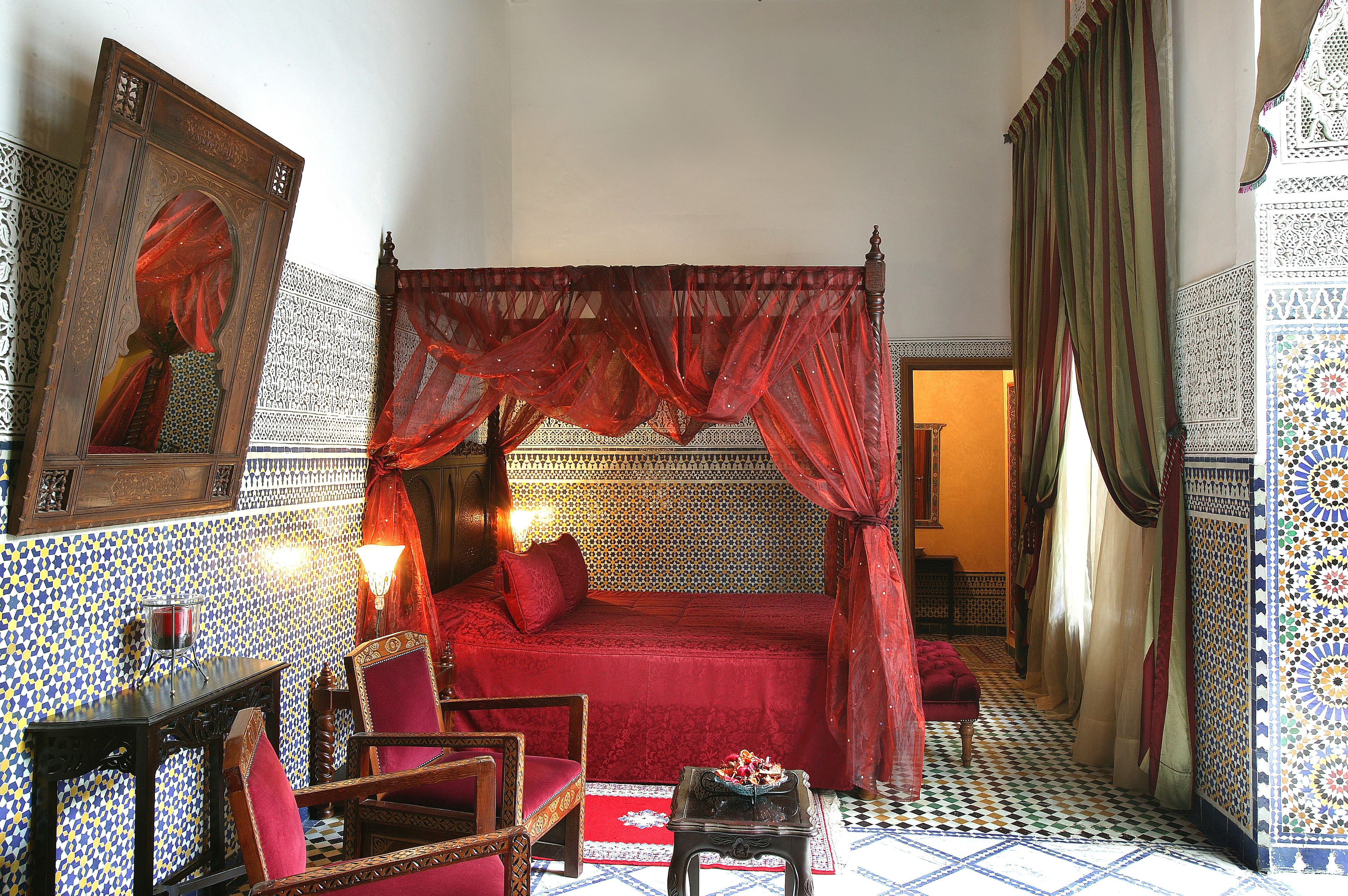 A room with a four-poster bed, a sitting area with a chair and table, and a mirror on the wall.