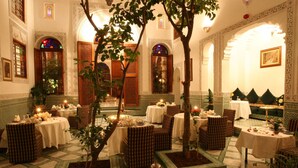 Un interior marroquí tradicional con arcos ornamentados, suelos de azulejos y un árbol en el centro. Se han dispuesto mesas para comer con velas.