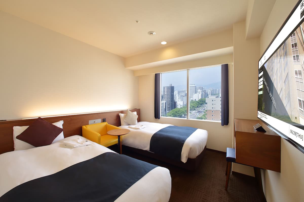 Habitación de hotel con dos camas, una mesita, una silla, televisión y ventana con vista a la ciudad.