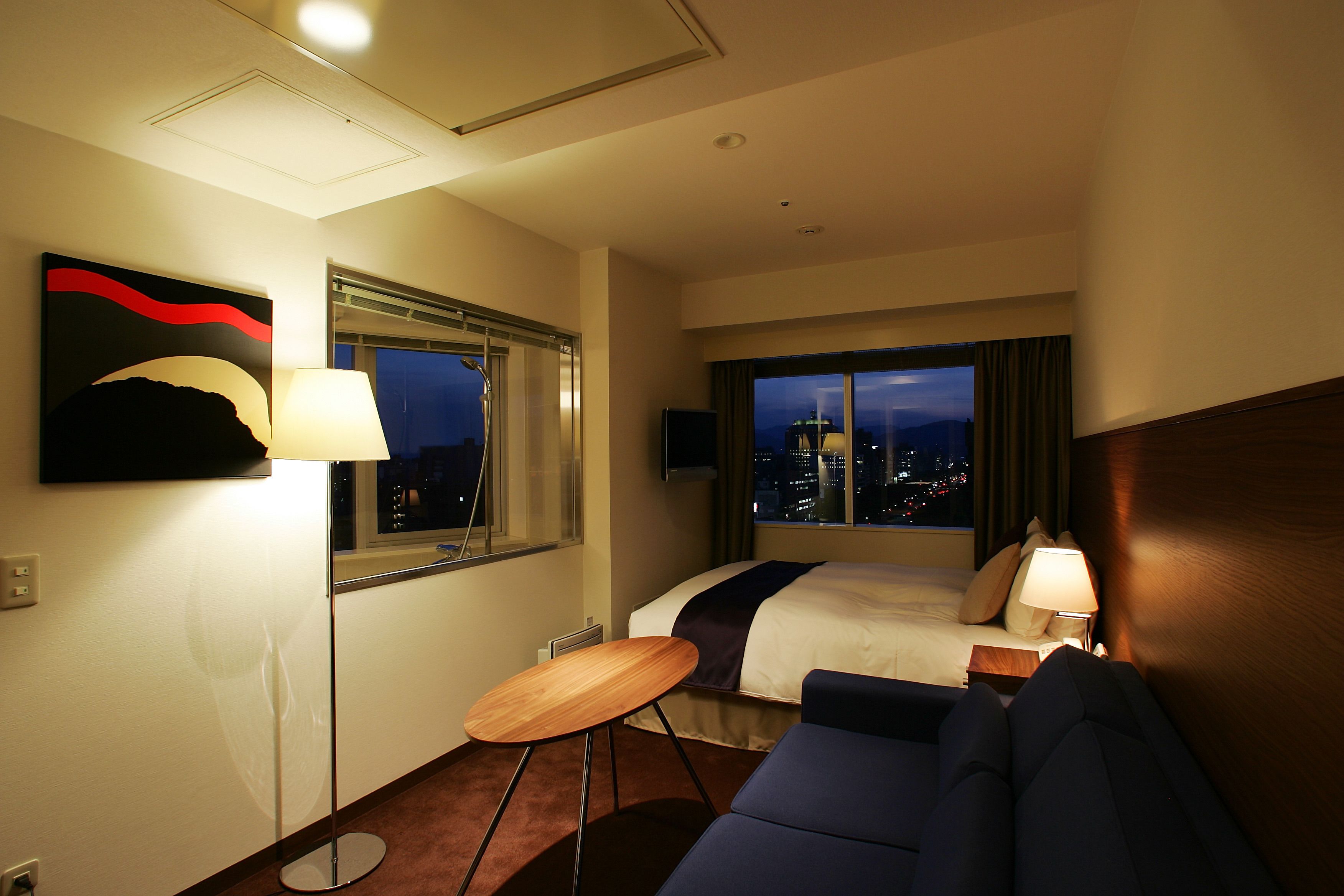 A hotel room with a bed, a desk, a chair, a lamp, and a large window with a city view.