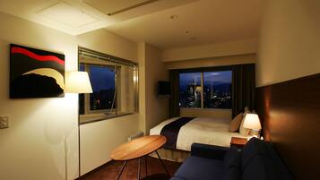 A hotel room with a bed, a desk, a chair, a lamp, and a large window with a city view.