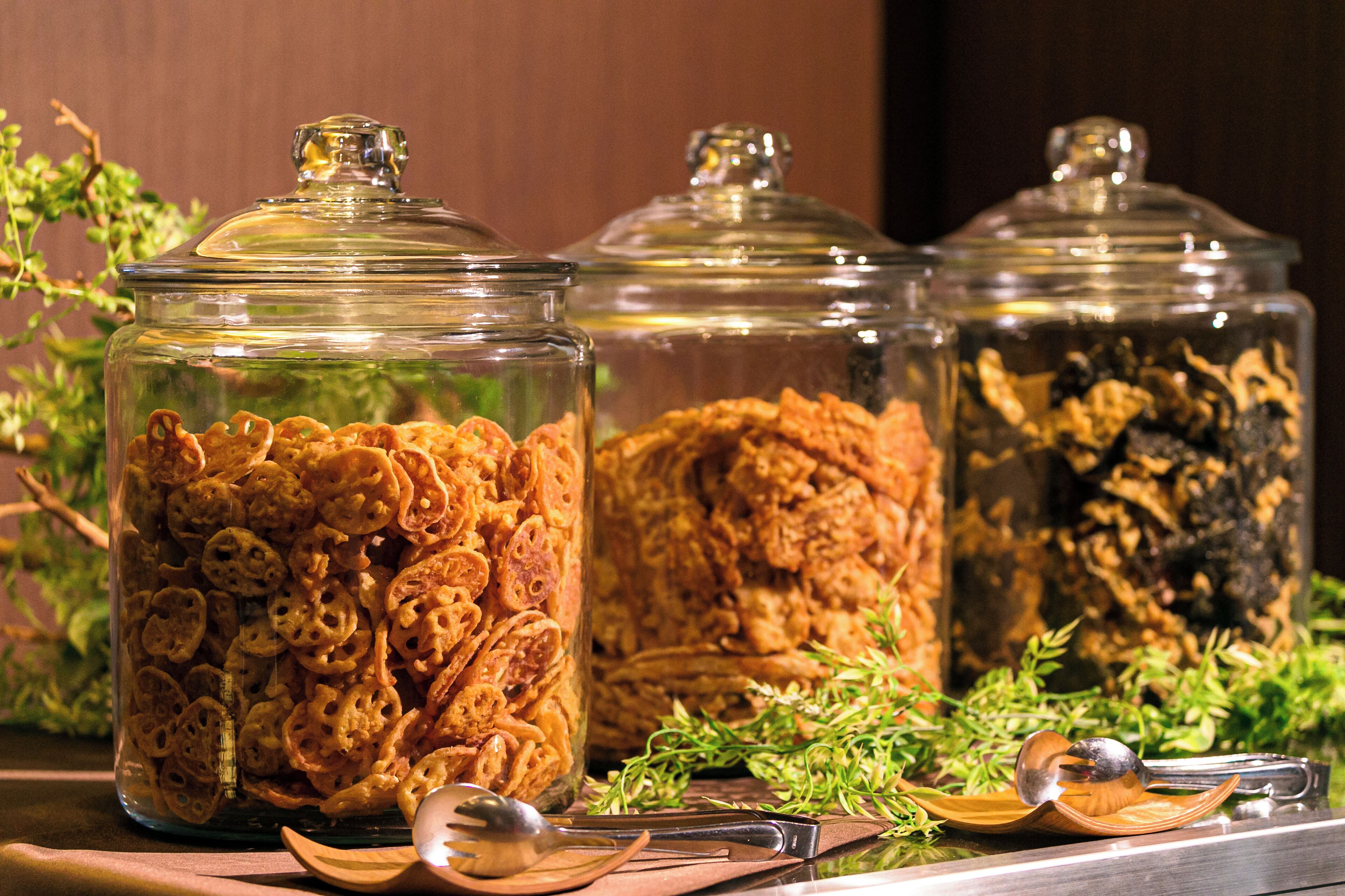 Trois bocaux en verre remplis de céréales sont posés sur une surface en bois, entourés de plantes et d’ustensiles de cuisine.
