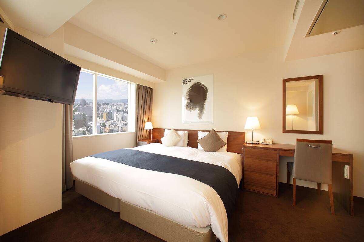 Un cuarto de hotel con una cama grande, un escritorio, una silla, un televisor, una ventana con vista a la ciudad y un cartel en la pared.