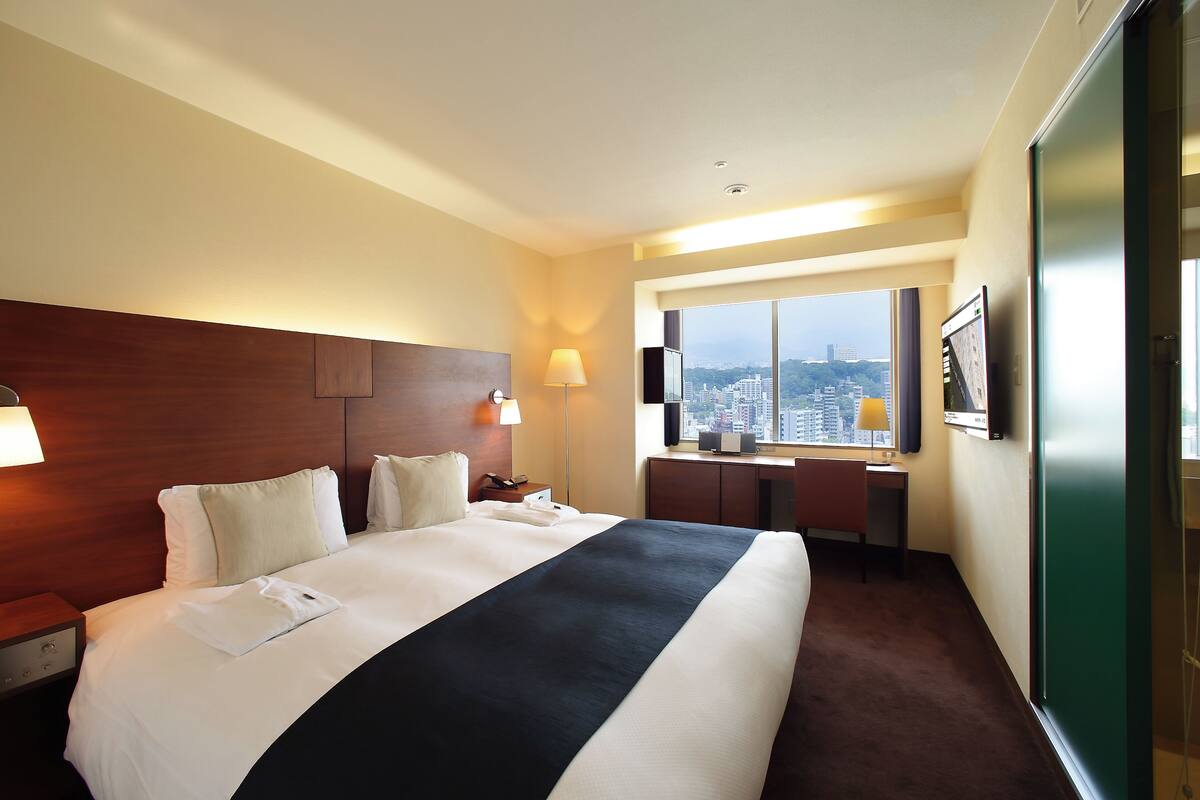 Habitación de hotel con una cama grande, un escritorio, una silla, un televisor y una ventana con vistas a la ciudad.