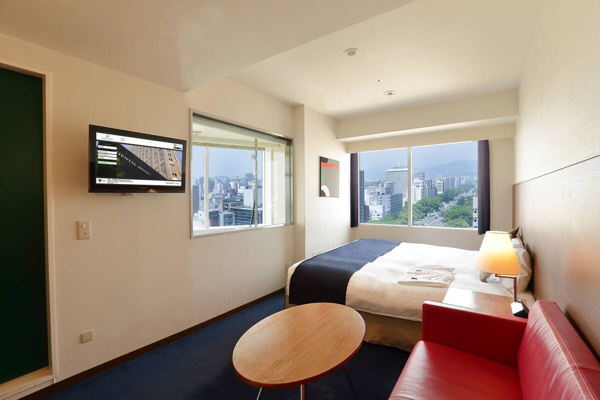 Habitación de hotel con cama, sofá rojo, una mesita, televisor montado en la pared y ventanas amplias con vista a la ciudad.