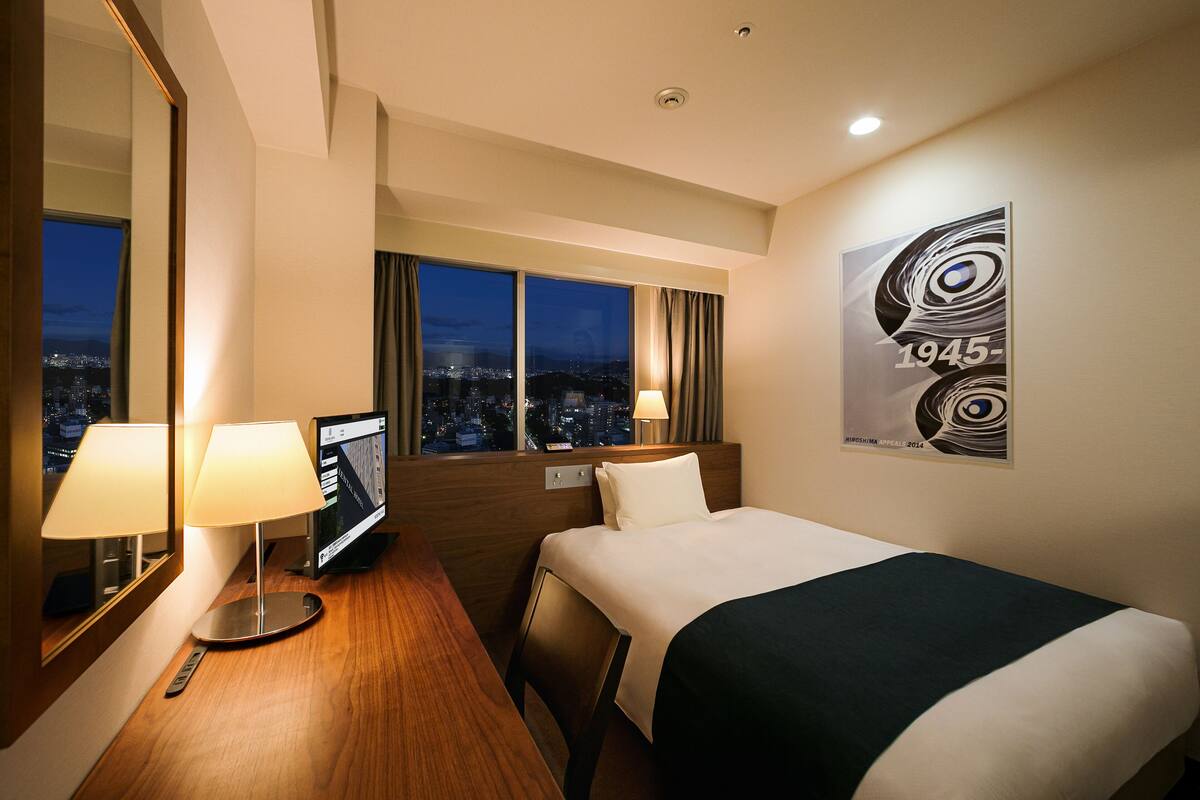Habitación de hotel con cama, dos lámparas de noche, televisor de pantalla plana, vistas urbanas a través de amplias ventanas y una obra de arte abstracta moderna en la pared.
