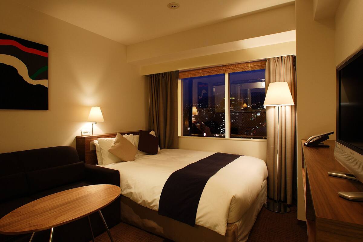 Habitación de hotel con cama, escritorio, silla, televisor y ventana con vista a la ciudad.