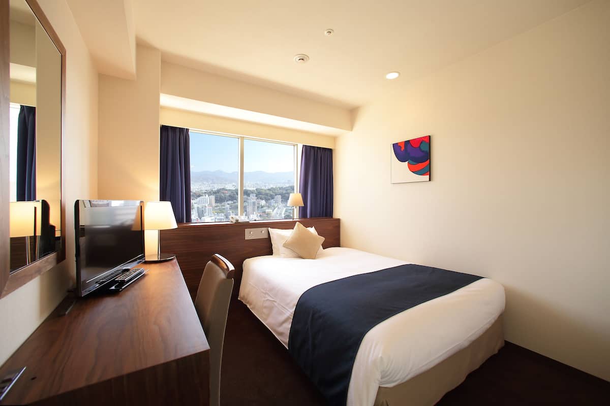 Habitación de hotel con cama, escritorio, silla y un amplio ventanal con vistas a la ciudad.